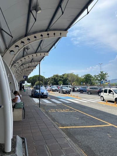 The image depicts a daytime scene at what appears to be an airport or train station in Lamezia Terme, Italy. The orientation of the image is vertical, with the sky occupying the upper portion and a parking lot filled with cars dominating the middle and right side. The left side features a modern, curved architectural structure, possibly a covered walkway or part of a terminal building, with lighting fixtures mounted on its underside.

Several cars are parked in a line along a road marked with yellow and white lines, indicating a designated parking area or crossing. Some of these vehicles are blue, white, and gray. A blue police car is prominently visible among the parked vehicles. In the foreground, on the right, a young girl with a white shirt and shorts is crouched down, possibly attending to something on the ground. A green backpack is placed next to her.

The sky is bright with scattered white clouds, suggesting a clear and sunny day. The presence of trees in the background indicates a relatively green environment. The architectural style of the building on the left, along with the presence of what looks like signage in the distance (though the text is illegible), points towards a transportation hub. The overall atmosphere appears calm and ordinary, with no obvious signs of urgency or distress. The time of day is clearly daytime due to the bright sunlight and clear skies.