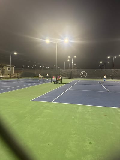 A tennis court at night is lit by several bright overhead lights. The court is green and blue. A net divides the court into two halves. There are people playing or practicing on the court. A ball machine is present. There is a building visible in the background. The sky is dark and unclear.