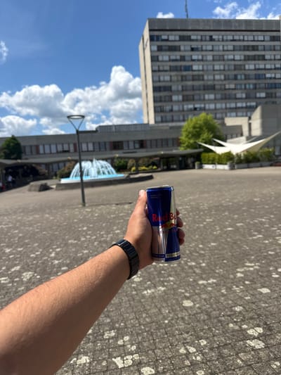 The image depicts a first-person perspective of a hand holding a can of Red Bull energy drink. The background reveals an outdoor urban setting. In the background, there is a modern architectural building with numerous windows, a public fountain, and various trees. The ground is paved with concrete stones. Based on architectural cues and the presence of the fountain, the location is likely near the Zentrumsplatz (Central Square) in Bottmingen, Switzerland. There are no other identifiable people in the image, only the arm and hand of the individual holding the can.