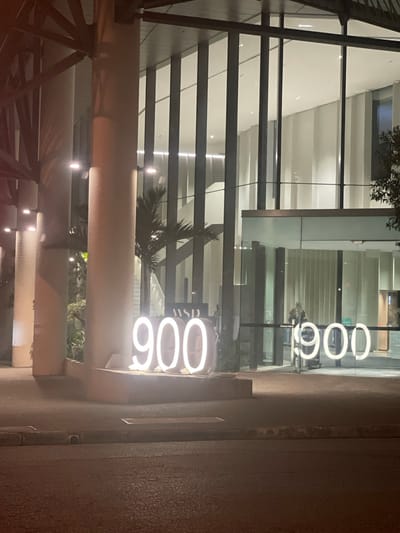 The image depicts the exterior of a modern building at night, with a focus on illuminated numbers displayed prominently. The building features a large glass facade, allowing views into a well-lit interior with vertical structural elements and a high ceiling. In front of the building, large, bright white neon numbers "900" are visible on the left side of the image, and another set of "900" numbers are visible on the right side, displayed on the glass. A palm tree is visible behind the "900" on the left, and another is partially visible on the right. A person is seen through the glass on the right, standing near the entrance. The street in front of the building is paved and appears wet, suggesting recent rain. The architectural style and the presence of tropical foliage, like the palm trees, are consistent with Brisbane, Australia. The lighting suggests it is either early morning or late evening.