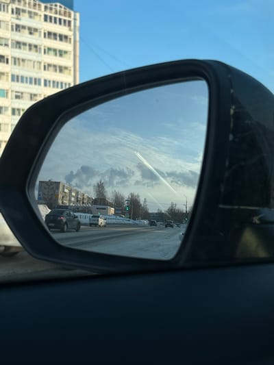 The image presents a view from inside a vehicle, focused on the driver's side mirror. Outside the upper-left of the mirror, a tall, light-colored multi-story residential building with numerous windows is partially visible against a clear blue sky.

The side mirror reflects a wide, snow-covered or icy urban road in Dobryanka, Russia, showing multiple lanes. Several vehicles are present on the road, including a dark sedan in the foreground of the reflection, and other cars further down the road, all appearing to travel in the same direction. To the left of the reflected road, a row of darker, multi-story apartment buildings with balconies and distinct architectural patterns are visible. Smoke plumes rise into the sky from behind these buildings. Bare deciduous trees line the road and intersperse with the buildings. A green traffic light is visible on the right side of the road, along with streetlights. The sky reflected in the mirror displays both blue areas and scattered clouds, with prominent white contrails extending diagonally across it. Snow accumulates on the road shoulders and median.