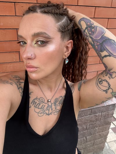 A close-up, rotated photograph shows a person with multiple tattoos and braided hair lying down. They are wearing a black, ribbed tank top. The person has green eyes, light skin, and wears subtle eye makeup. A silver necklace with a small, clear stone pendant is visible on their chest, over a tattoo. They also have a nose ring and an earring in their left ear, which is adorned with a teardrop-shaped, iridescent jewel. The background consists of reddish-orange brick tiles and a textured stone wall, suggesting an outdoor setting. The lighting appears to be natural, indicating daytime. The person's expression is neutral, with their head tilted back and eyes looking upwards. The tattoos on their arms are prominent, including a large purple octopus on their left arm and lettering on their chest.