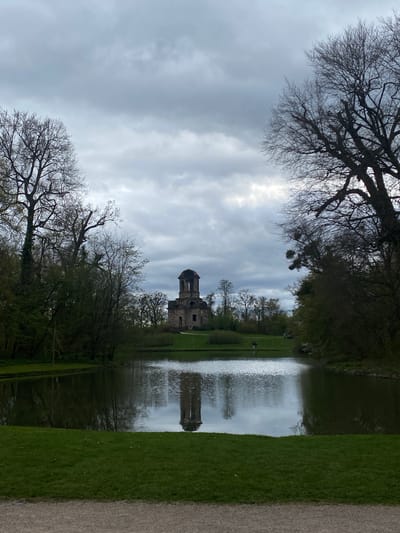 This image shows the ruined Temple of Mercury in the park of Schwetzingen Palace, Schwetzingen, Germany. The temple is reflected in the still water of a pond. Trees frame the view, some bare and some with fresh green leaves. The sky is overcast. There are a few figures of people in the distance in the green space near the pond. A pathway runs along the right side of the image.