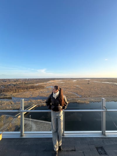 A sole female subject is observed standing on an elevated outdoor observation deck in Tårnby, Denmark, overlooking the natural landscape of Amager Fælled. She is positioned centrally, facing approximately 3/4 left, with her gaze directed towards the distant horizon. The subject has long, dark brown hair tied in a high ponytail. She is wearing a dark, possibly black, glossy puffer jacket, a light-colored (likely white or off-white) scarf, light blue wide-leg jeans, and dark shoes. Her hands are placed on her hips or tucked into her pockets.

The observation deck features a modern railing system composed of horizontal silver-colored bars and vertical support posts, with transparent panels (presumably glass) between them. The floor of the deck is a dark, tiled or paved surface, with a metal drain grate visible in the lower right corner. A metal chain is attached to a railing post on the right side.

The background reveals an expansive natural area characteristic of wetlands or marshland, dominated by light brown or tan dry vegetation, interspersed with numerous winding water channels and larger bodies of water reflecting the sky. In the far distance on the left, a discernible line of urban development, including buildings and trees, is visible. The sky is predominantly clear and bright blue, transitioning to a lighter hue with faint clouds near the horizon on the right, where the setting or rising sun casts a warm, soft light across the landscape and illuminates the subject from the right.