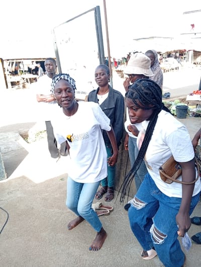 The image depicts a lively outdoor scene in Jebbu Miango, Nigeria, likely within a market or public thoroughfare. Six individuals of various ages are visible. In the foreground, a young woman with dark skin, wearing a white t-shirt with a multi-colored logo and light blue jeans, stands barefoot with her knees bent, a patterned headscarf on her head, and a direct smile towards the viewer. Her toenails are painted blue. To her right, another young woman with dark braided hair, wearing a white t-shirt, ripped dark blue jeans, and light sandals, is also in a bent-knee stance, looking down and to her right, with a brown shoulder bag.
Behind them, a young man in a white t-shirt stands to the left, looking away from the camera. A young woman in a dark grey jacket and light top stands between the foreground figures and the background, observing. Further back on the right, a person in a light bucket hat and another individual are partially obscured near market produce like green vegetables and a watermelon. In the far background, market stalls with corrugated roofs and wooden structures are visible. A large, white rectangular signboard with dark edges stands centrally in the mid-background. The ground is a light-colored paved surface, and various sandals are scattered near the feet of the individuals. A black cable runs from the bottom left corner across the pavement. The scene is brightly lit by natural daylight.