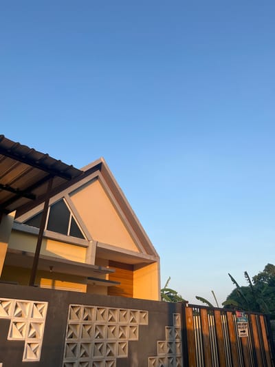 The image depicts a modern house in the city of Bandungrejo, Indonesia, bathed in the warm, low light of either sunrise or sunset. The clear blue sky takes up the majority of the frame, with a hint of green foliage visible on the right side. The house itself features a striking triangular gable, painted in a soft yellow, with a contrasting grey trim. A corrugated metal awning extends from the left side of the house, casting a shadow on the structure below. A grey concrete wall with decorative geometric openings runs across the foreground, separating the property from the street. To the right, a wooden gate with vertical slats stands partially open, and a sign that reads "DIJUAL" (For Sale) with a phone number is attached to it. The overall impression is one of a residential property in a tropical climate, likely in a suburban or semi-rural area, given the presence of banana trees and the clear, unobstructed sky. There are no people visible in the image.