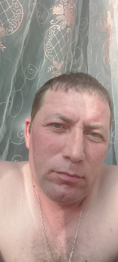 A close-up, low-angle selfie captures a middle-aged Caucasian man with short brown hair and blue eyes. He is shirtless and wearing a silver chain necklace. His face is flushed, with prominent redness on his cheeks and forehead, possibly indicating sunburn or a medical condition. His expression is neutral.
The background consists of sheer curtains with an embroidered pattern of floral elements and swirling motifs in a peachy-pink color against a pale green fabric. The lighting suggests it is daytime, possibly indoors near a window. No text is visible in the image. The location context is Абалак, Russia.