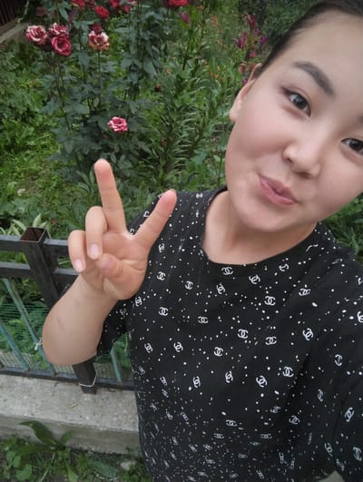 A young woman is taking a selfie in front of a rose bush. She is wearing a black t-shirt with a white pattern. Her right hand is held up in a peace sign. The background shows a rose bush with pink and red flowers, as well as some green foliage and a metal fence. The image appears to have been taken outdoors in a garden setting. No specific landmarks or features of Ак-Дөбө, Kyrgyzstan are visible.