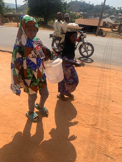 The image depicts a daytime scene in Egbedi, Nigeria, showing a partially unpaved environment adjacent to an asphalt road. In the foreground, two individuals, likely children, walk on a reddish-brown dirt path. The individual on the left, wearing a multi-colored patterned tunic and head covering, grey leggings, and light blue slides, holds a clear plastic Coca-Cola bottle in their right hand and a clear plastic lidded bucket in their left. This individual faces forward, making eye contact with the viewer. To their right, a second individual, wearing a dark head covering and a purple patterned dress with dark sandals, carries a small rectangular object in their right hand and walks towards the right of the frame, partially looking towards the viewer. Both individuals cast distinct shadows onto the dirt path. In the midground, on an asphalt road, a dark-colored motorcycle is in motion, carrying two adult males. Both males wear light-colored traditional attire and caps. The passenger, seated behind the driver, balances a flat, round tray containing several white, rounded objects on his head. The background features sparse green vegetation, hills, and a cluster of buildings with corrugated metal roofs, indicative of a rural settlement. The sky is bright and clear.