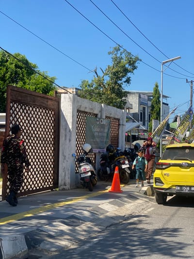 A sunny day in a city, likely in Jawa, Indonesia, is depicted. In the foreground, a person in a camouflage uniform stands near a large, ornate gate. Behind them, several motorcycles are parked along a sidewalk. A yellow car is parked on the street, with a speed bump in front of it. A young boy in a vest and shorts walks with an adult woman wearing a headscarf. Flags are planted along the street, some with text in Indonesian. A banner with text in green and white hangs on a wall. The environment includes a clear blue sky, trees, buildings, and overhead power lines.