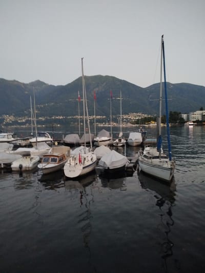 The image depicts a marina in Muralto, Switzerland, with several boats moored on a calm body of water. The sky is overcast, and the surrounding landscape features rolling hills and mountains. In the foreground, a white sailboat with a blue sail is docked. Other boats of various sizes, some covered with tarps, are also visible. The water reflects the boats and the overcast sky. The scene appears to be captured during twilight or early evening.