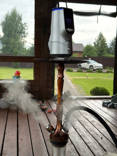 The image shows a shisha setup on a wooden deck, likely outdoors in a suburban or rural area. The shisha device has a silver and copper colored body with a black hose emitting smoke. Above the shisha, a silver electronic device with a screen displaying "84%" is visible. In the background, through a window or open area, a white car is parked on a green lawn with a fence and trees. There are also some children's toys, shoes, and other items scattered on the deck. The overall scene suggests a leisurely outdoor activity. The metadata indicates the location is Борисьево Днп, Russia.