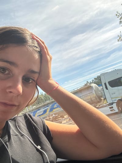 The image presents a close-up, selfie-style shot of an individual in an outdoor, industrial-leaning setting within Leiria, Portugal, under bright daylight.
**Person:** A single individual, appearing female, occupies the foreground. Their face, featuring brown hair and dark eyes, looks directly at the camera with a neutral expression. The right arm, displaying a tanned complexion and a pink string bracelet on the wrist, is bent, with the hand partially supporting the head. The individual wears a dark, ribbed top and white wired earbuds, with the cable visible across their chest.
**Subjects/Objects (Background):** The background reveals a bright blue sky with scattered white, wispy clouds. High in the sky, a faint grid of bright artificial lights is visible. Directly behind the individual, a large blue and yellow industrial structure or barrier is prominent, clearly displaying the "Cimentos Asland" logo. Adjacent to this, parked heavy vehicles include the cab of a white truck, marked "3720", and a silver cylindrical tanker trailer, labeled "4549". Below the vehicles, various stacked materials, possibly wood or construction beams, are visible. Green foliage from distant trees lines the far right background.
**Actions/Interactions:** The individual is posing for a photograph, with their head resting on their hand, while seemingly listening to audio via earbuds. No other individuals or direct interactions are depicted.
**Environment:** The scene is illuminated by strong, direct sunlight, indicating clear weather. The presence of large trucks, a tanker trailer, and an industrial structure with a cement company logo indicates an active transport or industrial zone.