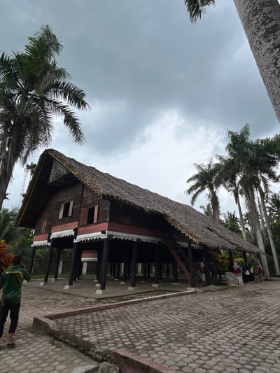 In the city of Mesjidpirak, Indonesia, an image captures a traditional stilt house with a thatched roof, surrounded by lush palm trees under a cloudy sky. The house, constructed of dark wood with red accents and white decorative trim, stands on numerous dark, slender pillars. A wooden staircase leads up to an open entrance on the side of the house. The ground around the house is paved with hexagonal interlocking stones, bordered by a low, reddish-pink concrete curb. A person in a green shirt and dark pants is seen walking away from the camera on the left. Several other individuals are standing near the house on the right, some appearing to be tourists or visitors. The overall scene depicts a cultural heritage site within a tropical environment.
