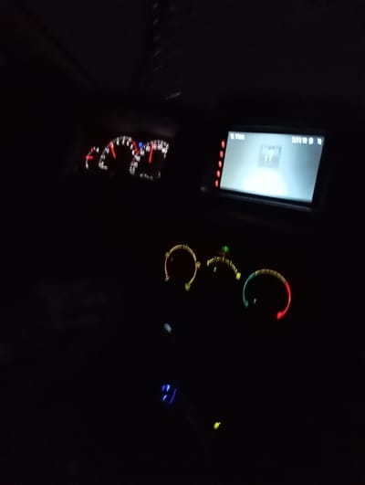 The image depicts the interior of a car at night, likely in Krasnoyarsk, Russia, given the location context. The dashboard is visible, illuminated by the car's internal lights. A speedometer and tachometer are partially visible on the left, showing readings in the red zone for speed, indicating the car is in motion. A multimedia screen is centered in the dashboard, displaying a bright, almost blank white screen with some faint text at the top. Below the screen are circular control knobs for what appear to be climate control or other vehicle functions, some of which are illuminated with colored LEDs (yellow, green, and red). The overall environment is dark, with the only significant light sources being the dashboard instruments and the screen. There are no people visible in the image. The scene captures a moment of driving at night, emphasizing the illuminated controls and displays of the car's interior.