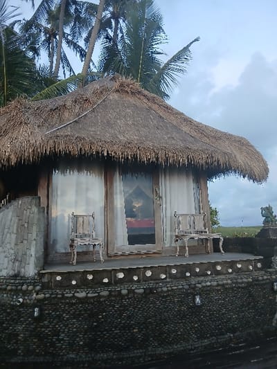 This image depicts a traditional-style bungalow located in Tibubiyu, Indonesia. The structure features a prominent conical roof constructed from dried thatch. Below the roofline, the facade includes a central wooden-framed glass door, flanked by two similarly framed glass windows. Internal white curtains are partially visible behind the glass. Reflections of a sunset, displaying orange, red, and purple tones against a cloudy sky, are visible on the glass surfaces.
A wooden deck extends across the front of the bungalow. Two light-colored, ornately carved wooden chairs are positioned on this deck, one to the left and one to the right of the central door. The dwelling is supported by a multi-tiered foundation. The upper section of the foundation is a dark surface with evenly spaced circular depressions, while the lower, more expansive section is covered entirely with dark, smooth river stones. Two dark, lantern-style outdoor light fixtures are affixed to this stone-clad foundation.
To the immediate right of the bungalow, a dark stone wall or railing is present, topped by a carved stone statue resembling Ganesha. In the background, multiple tall palm trees with green fronds rise against a light blue-grey, overcast sky. The distant environment beyond the stone wall appears to be an open, flat landscape. No individuals are present in the scene.