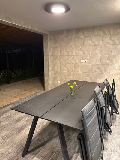 The image depicts a covered outdoor dining area, likely a veranda or patio, in Peyovstsi, Bulgaria, illuminated at night. A circular, flush-mounted LED light fixture on the white ceiling provides primary illumination. The right wall is covered in light-beige, rectangular, rough-textured tiles, with a white electrical outlet and switch panel positioned centrally. The floor is tiled with elongated planks resembling dark grey-brown wood grain.
A long, rectangular outdoor dining table, dark grey with a textured surface and dark angled legs, occupies the center-right of the frame. Four dark grey metal-framed folding chairs with mesh seats and backs are positioned along the right side of the table, appearing stacked or partially folded. A small, clear glass vase containing three yellow daffodils sits on the table, slightly to the left of its center line.
To the left, a large open entryway leads to a dimly lit exterior. The outdoor area features light-colored square paving stones and dark green low-lying vegetation. A darker wooden or metal structure is partially visible in the distant background. No individuals or active interactions are present within the scene.