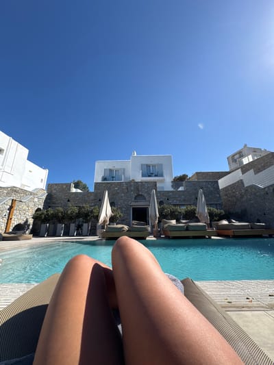 The image depicts a first-person perspective of a poolside area in Mykonos, Greece. In the foreground, two tanned human lower legs rest on a woven sun lounger, with a small section of a grey textile visible near the left leg. A clear, blue swimming pool occupies the midground. Beyond the pool, a terrace features several daybeds with wooden platforms and grey cushions, interspersed with three large, light-colored, folded beach umbrellas. Green plants in rectangular planters line the base of a prominent stone retaining wall. In the background, traditional Cycladic architecture is present, characterized by whitewashed buildings with light blue window shutters, including a two-story structure with two balconies. Other similar buildings are visible on the periphery and higher on a distant hillside. The sky is uniformly clear and bright blue, with direct sunlight casting sharp shadows and producing a lens flare in the upper right.