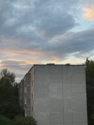 The image captures a view through a window screen, overlooking an apartment building in Pinsk, Belarus. The building appears to be a multi-story, concrete structure with visible windows and a flat roof with several protruding structures. Trees and foliage surround the building on the left and right sides, suggesting a residential area. The sky above is filled with clouds, with hints of orange and yellow, indicating either sunrise or sunset. The overall scene is residential and calm, with no people or specific actions visible. The perspective suggests the photograph was taken from another building or a higher vantage point.