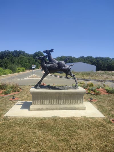 The image shows a bronze sculpture of two deer, one positioned above and slightly behind the other, in mid-gallop. The sculpture is mounted on a rectangular, fluted pedestal, which rests on a concrete slab. This monument is situated outdoors in a landscaped area with dry grass, gravel, and some low-lying plants.
In the background, a paved road curves to the left. A road sign, "E15 LESNEVEN-METZ," is visible near the road intersection. Beyond the road, there is a line of lush green trees forming a dense forest. A large, light-colored, corrugated metal building with a sloped roof stands behind the trees on the right side of the frame. The sky above is a clear, bright blue. The scene suggests a rural or semi-rural environment, possibly at the edge of a town or near a highway, in Ressons-sur-Matz, France. The lighting indicates a sunny day. There are no people or animals present other than the sculpture.