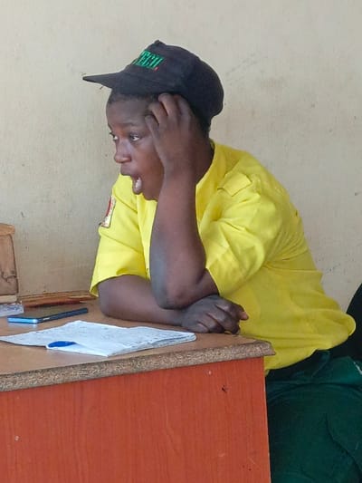 The image captures a close-up of a person seated at a wooden desk, seen from the chest up, positioned slightly to the right of the frame, facing left. The person appears to be a young adult with dark skin. They are wearing a black baseball cap with a green logo that partially reads "TECI" or similar green lettering on the front. Their facial expression shows a wide-open mouth, as if in surprise, shock, or a yawn, with their eyes looking intensely towards the left, out of frame. One hand is raised, with fingers touching or scratching their head near the temple, suggesting either distress, deep thought, or weariness.
The person is wearing a short-sleeved, bright yellow collared shirt. A small patch or emblem, predominantly red and white, is visible on their left shoulder. Below the desk, a glimpse of dark green fabric, possibly pants or a skirt, can be seen.
On the light brown wooden desk in front of them, there are several items. A dark blue or black smartphone is lying flat on the desk, along with a stack of white papers or a notebook, on which a blue pen is resting. The desk itself has a speckled texture on its surface.
The background is a plain, light-colored wall, possibly off-white or light beige, which appears somewhat textured. The lighting suggests an indoor setting, with no clear indication of the time of day. The overall impression is one of candidness, capturing a moment of strong reaction or emotion from the individual.