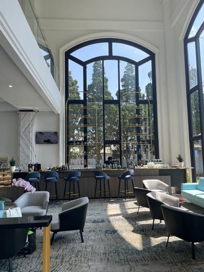This image depicts a modern, high-ceilinged interior lounge area, possibly within a hotel or café, located in Kayuambon, Indonesia. The scene is dominated by two large, tall, black-framed arched windows on the right and center, providing ample natural light and an exterior view.

In the foreground, a patterned carpet in shades of grey, blue, and brown covers the floor, exhibiting distinct shadows cast by sunlight entering the windows. Seating arrangements include a black rectangular table with a golden vertical support on the left, beside which a white handbag rests and a black reusable bag is on the floor. Four armchairs are visible: two light grey upholstered chairs with black legs and two darker grey/black upholstered chairs with black legs, positioned facing away from the viewer. A light blue sofa is partially visible on the far right. Pink orchid flowers extend into the frame from the extreme left.

The midground features a long, dark wood bar counter with a lighter front panel. Four dark blue upholstered bar stools with black legs and golden footrests are arranged along the counter. Behind the counter on the left, a white marble-patterned pillar is visible next to a white wall with a mounted flat-screen television displaying an indistinct image. Bottles of spirits are arranged on the bar counter, alongside smaller plants and decorative items. A sign partially reading "GELATO" and "KURT COBAIN" is visible on a lower shelf. In the center, a prominent golden arch-shaped shelving unit stands in front of the main window, displaying various glasses and bottles. To the right of the shelving unit, coffee cups and brewing equipment are visible on the counter. One person, a male in a dark shirt, is seated behind the bar, facing the counter, with their face partially obscured.

The background is dominated by the large windows, which reveal dense green coniferous trees and a clear blue sky outside. Through the window on the far right, a blue "EXIT PARKING" sign is visible, along with a glimpse of a building featuring a red roof. Above the main lounge area, a white mezzanine level with a clear glass railing is visible on the upper left, while white architectural molding and columns adorn the walls around the windows, contributing to the elevated ceiling design.