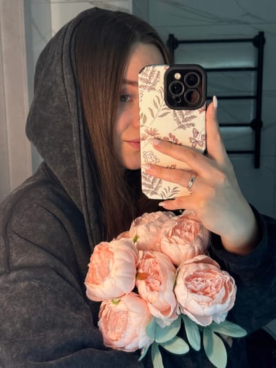 A female individual is depicted taking a mirror selfie in a domestic setting within the city of Tliustenkhabl', Russia. She is partially visible, with her face mostly obscured by a light-colored smartphone featuring a botanical pattern case, held in her left hand. Her right eye and the right side of her face are visible, along with her long, dark brown hair. She is wearing a dark gray, faded hooded sweatshirt. Her left hand displays manicured nails with light polish and a ring on the ring finger. In her right arm, she holds a bouquet of approximately six light pink, artificial peonies with green artificial leaves. The background, reflected in the mirror, shows a light-colored tiled wall and a black, wall-mounted heated towel rack.