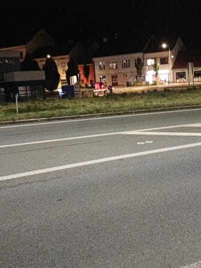 The image depicts a nighttime scene on a road in Nový Jičín, Czechia. In the background, there is a row of buildings with illuminated windows, suggesting commercial or residential use. A street light is visible, casting a glow. In the middle ground, there is a grassy area and a dark structure that appears to be a building or kiosk. A blue bin or container is also present. The foreground shows a paved road with white lane markings. The overall impression is that of a quiet urban or suburban street at night.