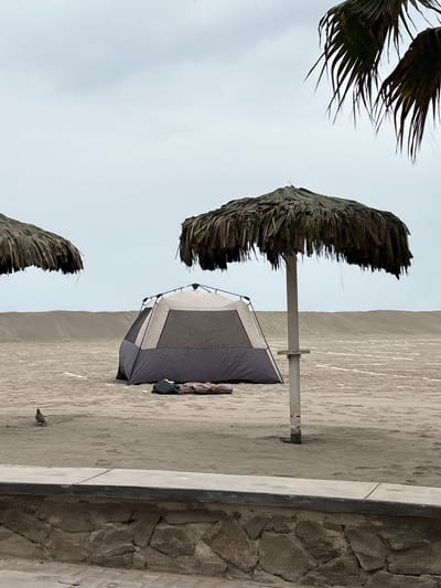 This image depicts a serene beach scene in the city of San Antonio, Peru, under a heavily overcast sky. The foreground features a rough stone wall topped with a concrete ledge. Beyond the wall, a sandy beach stretches out, marked by faint footprints and some scattered debris. A small bird stands on the sand to the left of the frame.
Dominating the mid-ground is a large, two-tone tent in shades of gray and tan, set up on the sand. In front of the tent, there are some bags and rolled-up bedding, suggesting an overnight stay. To the right of the tent, a large palapa umbrella with a thatched roof stands on a pole, providing shade. Another, similar palapa umbrella is partially visible on the far left of the image. In the background, sand dunes rise up, creating a natural barrier. The overall atmosphere is one of quiet solitude, with the muted colors of the sky and landscape contributing to a tranquil mood. There are no people visible in the image.