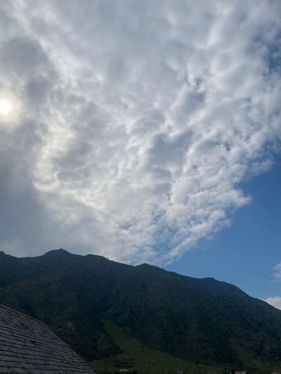The image shows a mountain range under a partly cloudy sky. A sun is visible on the left side of the frame, partially obscured by clouds. In the foreground, the roof of a building made of wooden shingles is visible. The mountain slopes are covered in vegetation, with a few scattered trees visible on the ridges. The sky is a mix of blue sky and white and gray clouds, with the clouds appearing denser and darker on the left side of the frame. The overall impression is one of a natural landscape, possibly a rural or mountainous area. The provided crops indicate that this image might be from Inya, Russia, suggesting a mountainous and forested region. However, there are no specific landmarks or identifiable features that would pinpoint the exact location within Inya, Russia. There are no people or animals visible in the image. The lighting suggests it is daytime, with the sun's position indicating it might be morning or late afternoon.