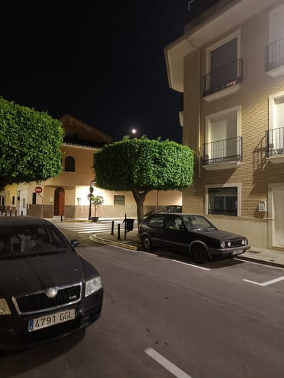 This is a night-time, medium-shot, eye-level, outdoor photograph of a residential street in Benirredrà, Spain. The foreground shows the front of a dark-colored Skoda Octavia with its license plate visible as "4791 GGL". To the right of the Skoda, parked on the street, is a dark-colored Volkswagen Golf. The street has white lane markings and a crosswalk. In the background, there is a building with a light-colored brick facade and balconies. Two large, round, manicured trees are positioned on either side of the street in front of a beige building. A full moon is visible in the dark night sky. The scene appears to be quiet and empty of people.