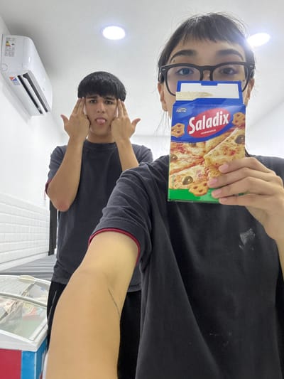Two people pose in an indoor setting. The person in the foreground wears a dark shirt with red trim and holds a "Saladix" cracker box over their eyes. The background person, wearing a similar dark shirt, sticks their tongue out while holding their hands near their cheeks. An air conditioning unit is visible on the left side of the image, positioned on a white wall with round ceiling lights. A portion of what looks like a tiled surface and a brightly colored bin are visible at the bottom right.