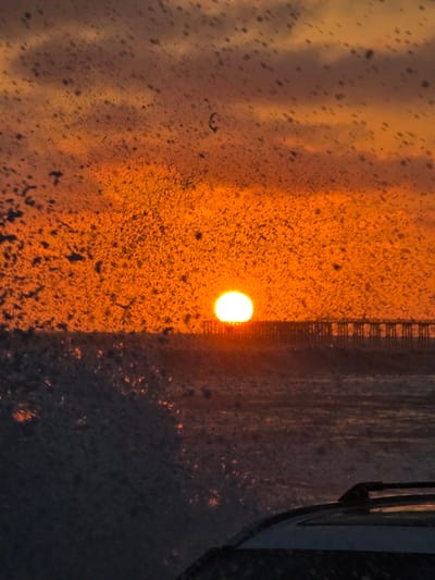 A setting sun casts an orange glow over the ocean in Oceanside, California. The sun is a bright circle low on the horizon, peeking out from behind a distant pier. The water is dark and choppy, with whitecaps visible. The foreground is obscured by what appears to be a car windshield covered in water droplets, creating a speckled effect. Part of a car's roof rack is visible at the bottom right of the frame. The sky is a mix of orange and purple, with clouds forming streaks of color.