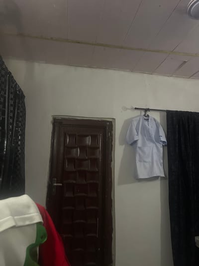 A medium shot captures the interior of a room, focusing on a dark brown wooden door with a metal handle, a white lab coat hanging on a hook against a pale wall, and a glimpse of a black curtain. The ceiling, paneled with light-colored wood, is visible at the top of the frame. To the left of the door, a black, patterned metal structure is partially visible, and in the foreground, on the left side of the image, a white garment with a green and red element is partially visible, suggesting it is draped over something or lying on the floor. The overall impression is that of a simple, functional space, possibly a bedroom or a storage area, located in Pantinapu, Nigeria.