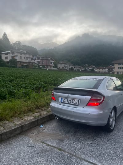 The image captures an outdoor scene in Elhovets, Bulgaria, on an overcast and foggy day, with low-lying clouds partially obscuring the forested mountains in the background. In the foreground, a silver Mercedes-Benz C-Class SportCoupé (C220 CDI model) with a black rear spoiler and a white sticker across its rear window is parked on the right side of an asphalt road, facing away from the viewer. The car displays a Bulgarian license plate, K.1214.BB, and its left rear brake light is illuminated red. The vehicle's rear bumper is equipped with parking sensors, and a dual exhaust tip is visible on its right side.

To the left of the asphalt road, a concrete curb borders a strip of grass, beyond which lies an extensive green agricultural field populated with dense, leafy plants, likely a crop such as potatoes. Further up the hillside, several multi-story residential houses of varying architectural styles and colors are visible, nestled among trees. These structures are situated at the base of the densely forested mountains, which disappear into the thick fog above. The sky is uniformly gray, and the overall lighting is diffused. No people are visible in the scene. On the asphalt surface near the curb, a blue wrapper and a slender dark object are present.