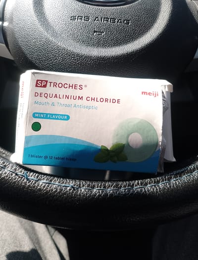 A retail box of SP Troches Dequalinium Chloride Mouth & Throat Antiseptic lozenges, mint flavor, is placed on a black steering wheel. The box is white with red and blue text. A green lozenge with a hole in the center, resembling a ring, is depicted on the front of the box, along with a few green mint leaves. Text on the box indicates "1 blister @ 12 tablet hisap." The "meiji" logo is visible on the top right of the box. The steering wheel appears to be in a car, with stitching visible along the rim. The overall lighting suggests it is daytime. The image does not provide any specific information about the location in the city of Meunasahuket, Indonesia.