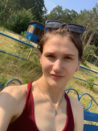 This is a portrait of a young woman outdoors, possibly taken during a sunny day. She is wearing a maroon top and a necklace with a cross pendant. Her hair is dark and short, and she has on large, dark sunglasses perched on her head. She appears to be smiling faintly, with a relaxed and content expression.
The background suggests a rural or natural setting. Behind her, there are trees, tall dry grass, and a small, blue wooden structure with a window. A blue metal railing, possibly part of a park bench or playground equipment, is visible in the foreground and along the right side of the frame. The lighting and the dry grass suggest it might be late summer or early autumn. The presence of the railing and the simple structure hints at a recreational area. There is no visible text in the image.