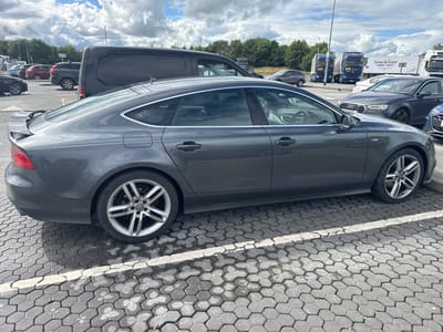 This media file captures a scene at a parking lot in Commons Cross, Ireland. The primary subject is a dark gray Audi A7 parked on a honeycomb-patterned paved surface. The car is seen from a rear-three-quarter view, showcasing its sleek design, including its distinctive alloy wheels and a subtle spoiler.

Other vehicles are visible in the background, suggesting a busy location. These include a dark-colored van, a red SUV, and several trucks, one of which has "Cuisine de France" written on its trailer. The setting appears to be an outdoor public space, likely a service station or a large parking area, given the presence of multiple large vehicles.

The weather seems partly cloudy, with a mix of sun and overcast skies, indicated by the shadows cast on the ground and the illuminated clouds in the sky. The time of day appears to be daytime, possibly late morning or afternoon. There are no people clearly visible in the frame, but the presence of parked cars and trucks implies human activity associated with travel or commerce. The overall impression is that of a typical, functional parking area.