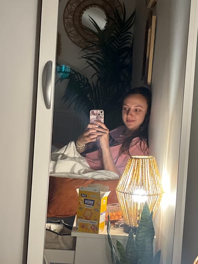 The image is a mirror selfie capturing a young woman in a room, holding her smartphone. She has long, dark hair and is wearing a pink top, with a bracelet visible on her wrist. Her expression is a soft, relaxed smile as she looks towards the phone, which has a light-colored, patterned case.
The reflection reveals a cozy interior. On the left, a large, dark green, palm-like plant stands prominently, partially obscuring a circular decorative mirror with a woven frame. Above the plant, wooden shelves are visible. To the right, a square-shaped lamp with a woven, straw-like shade emits a bright, warm light. Next to the lamp, an open yellow box of snacks, identified as "КУКУРУЗНЫЕ ЧИПСЫ" (Corn Chips) from the visible text, and a clear container with orange, round snack items are placed on a surface. Another green plant, possibly a snake plant, is partially seen near the snacks.
Elements of a bed or couch, including a rust-colored pillow and a white blanket, are visible. The bottom edge of the mirror appears to be part of a white piece of furniture with a silver handle. The warm lighting from the lamp suggests it might be evening or indoors. The scene is located in Vitry-sur-Seine, France.