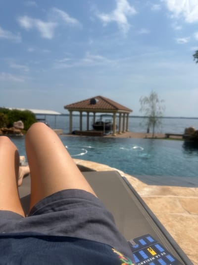 The image depicts an outdoor scene at a waterfront property in Montgomery, United States. The perspective is first-person, showing the lower legs and thighs of an individual, tanned and wearing dark blue shorts, resting on a dark gray lounge chair in the foreground. Beyond the person and chair, an infinity pool with blue water and visible surface ripples extends towards a large body of water, likely a lake or wide river. A tiled or paved patio area surrounds the pool.
In the mid-ground, a prominent boathouse or gazebo structure stands on the water. It features a reddish-brown tiled roof supported by numerous light-colored columns, with a dark-colored boat partially visible inside. To the left of this structure, a simpler, white-roofed dock or boathouse is also present on the water. The background consists of the expansive body of water under a clear blue sky dotted with scattered white clouds. Green foliage is visible on the left shoreline, and a slender, light-colored tree stands on the right shore. A colorful graphic displaying the text "DE LA MEDIANO" is visible on an item near the bottom right edge of the frame, adjacent to the person's leg. The scene is illuminated by bright sunlight, indicative of a clear day.