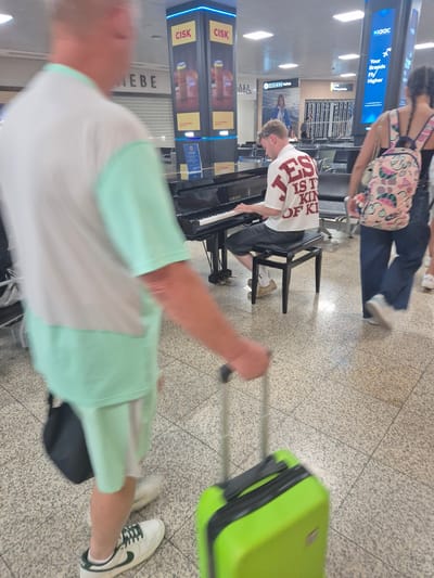 The photograph shows a young man playing a grand piano in a public space, likely an airport terminal in Luqa, Malta, given the context. He is seated on a black stool, wearing a white t-shirt with "JES IS TH KIN OF KI" printed in red lettering. He is wearing dark shorts and light-colored shoes. In the foreground, a person is walking away from the camera, pulling a bright green suitcase. This individual is wearing a white t-shirt with light green sleeves and shorts. Another person, a woman with a patterned backpack, is walking past the piano player in the background. The environment features tiled flooring and a large cylindrical advertising pillar with "CISK" branding. The overall scene captures a moment of impromptu musical performance in a travel hub.