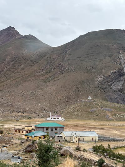 In Reru, India, a valley setting features a collection of buildings nestled at the base of imposing, barren mountains. The foreground shows a cluster of houses with yellow and green roofs, some with visible windows and doors. Behind these, a larger grey building with a green roof is prominent, alongside smaller structures. A dirt road winds through the area, and a vehicle is parked in the distance. The mid-ground displays a Buddhist stupa situated on a hill, with white lettering spelling out "JLMS STUPA" visible on the mountainside above it. The mountains are predominantly brown and rocky, with sparse vegetation in places, and are partially obscured by an overcast sky. The overall scene depicts a remote, high-altitude settlement against a dramatic mountainous backdrop.