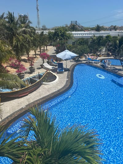 The image presents a bright, sunlit overhead view of a resort or hotel pool area, captured from a slightly elevated perspective.
The dominant feature is a large, irregularly shaped swimming pool, which occupies the right and lower-middle sections of the frame. The pool is tiled with a vibrant mosaic pattern of various shades of blue, giving the water a sparkling, inviting appearance. The water itself looks clear and clean, and a white inflatable ring floats near the top curve of the pool. A dark, narrow border surrounds the pool, separating it from a light grey paved path.
Along the paved path, several sun loungers are visible. In the mid-left portion of the image, a light blue umbrella shades one of these loungers, where a person can be seen relaxing in a blue swimsuit. Another person is visible further up on a different sun lounger.
A notable decorative feature is an old wooden boat, painted in weathered shades of blue and brown, which has been repurposed as a pond or planter. It's situated to the left of the pool, set into the landscaping, and contains water with what appear to be lily pads or other aquatic plants.
Lush green vegetation is abundant throughout the scene. Numerous palm trees are scattered across the left side of the frame and also frame the foreground on the bottom right. Bushes with clusters of bright pink flowers add splashes of color near the boat and sun loungers.
In the background, on the far left, several white, multi-story buildings with flat roofs are visible, suggesting resort accommodations. Beyond these structures, a tall, slender metal lattice tower, possibly a telecommunications mast, stands against the clear blue sky. Utility lines are also faintly visible in the upper left background.
The scene is bathed in strong, natural sunlight, indicating a clear and warm day, likely mid-morning or early afternoon, with distinct shadows cast by the trees and structures. The overall atmosphere conveys a sense of tropical relaxation and leisure.
A small black marker with the text "1m" is visible on the grey path next to the pool, indicating the pool's depth at that point.