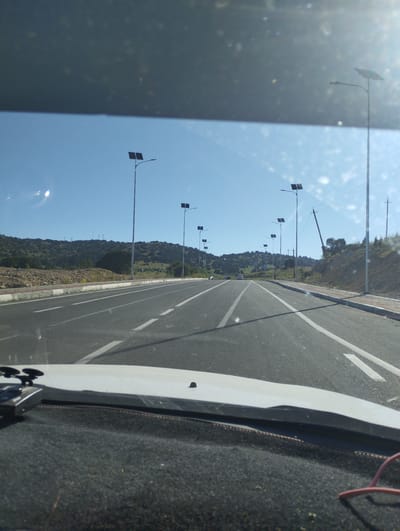 This image depicts a road in a rural or suburban area, likely on the outskirts of Myk, Uzbekistan, characterized by a clear blue sky and distant rolling hills covered with vegetation. The road itself is paved and appears to be a multi-lane highway with white lane markings. Along the side of the road, there are solar-powered streetlights, suggesting modern infrastructure development. The terrain is uneven, with some rocky and grassy slopes. There are a few cars visible on the road in the distance, indicating moderate traffic. The foreground shows the dashboard of a vehicle, with a device mounted on the windshield, suggesting the image was taken from the perspective of a driver. The overall impression is one of a well-maintained road leading through a natural landscape, possibly a developing area on the edge of a city.