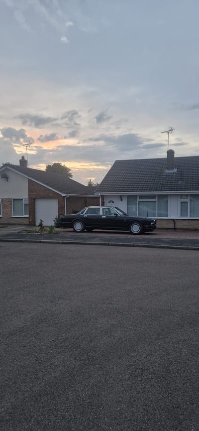 A black Jaguar XJ is parked on a residential street in South Holland, United Kingdom. The car is positioned in front of two detached houses. The house on the left has a white garage door and a brown brick lower half with a cream-colored upper half. The house on the right has a white front door and a beige brick lower half with a cream-colored upper half. Both houses have tiled roofs and white-framed windows. The sky above is overcast with some pale orange and pink hues visible, suggesting it is either dawn or dusk. The foreground consists of a tarmac road.