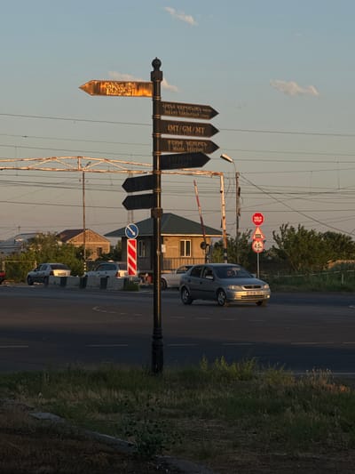 A street signpost with multiple directional arrows stands prominently on a road in Masis, Armenia. The signpost is black with gold lettering, pointing in various directions with text that appears to be in Armenian and English. The top arrow reads "BRANT VARDANYAN STREET," with Armenian text above it. The second arrow from the top says "IMT/GM/MT." The third arrow reads "MASIS STATION" with Armenian text above it. In the background, a railroad crossing with a red and white barrier is visible, along with a small building and houses. A silver sedan is driving on the road, and other cars are parked on the side. The scene is set against a clear sky with a few wispy clouds, suggesting it is daytime with the sun setting or rising. The environment is urban, with power lines and streetlights present.