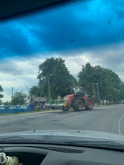The image shows a red tractor with a harvesting attachment on a road in Слабада-Кучынскі сельскі Савет, Belarus. The tractor is moving on the right side of the road, with trees lining the background. The sky is cloudy, and the overall lighting suggests it is daytime. A blue fence with a white decorative pattern runs along the side of the road. A small building with a gray roof is visible behind the fence, and some graffiti is present on the building's wall. The foreground shows the dashboard and windshield of a vehicle, indicating the image was taken from inside a car. The windshield is slightly dirty.