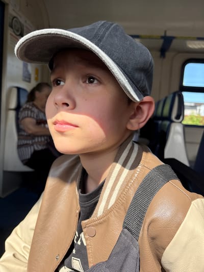 Inside a public transport vehicle, likely a train or bus, in Vyatskiye Polyany, Russia, a young male is prominently featured in the foreground, illuminated by strong sunlight entering from a window to his right. He is positioned facing right and looking slightly upwards, with the left side of his face bathed in direct light, creating a strong contrast and casting a shadow from his cap's brim over his upper face. He wears a dark grey baseball cap with a white brim, a light brown leather-like bomber jacket with off-white sleeves and striped brown and white cuffs/collar, and a dark t-shirt with a light graphic beneath. A dark grey strap, possibly from a bag, crosses his right shoulder. His light brown eyes are open, and his lips are slightly parted. In the blurred background, an adult female with medium-length hair, wearing a patterned top, is seated to the left, facing away from the camera. The vehicle's interior features light-colored walls, overhead grab bars with blue and yellow sections, and seats with blue and green striped upholstery and white headrest covers. A window on the right reveals a blurred outdoor scene with blue sky and green elements, indicative of daytime.