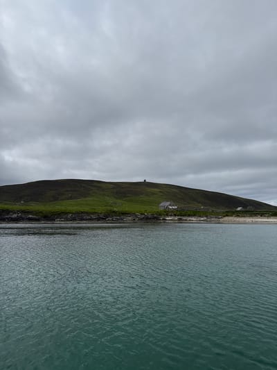 The image shows a body of water in the foreground and a green, rolling hill with some scattered buildings in the background. The sky is overcast with gray clouds. The water is a dark teal color and appears to be choppy. The hill is covered in lush green grass and appears to be uninhabited except for a few small structures. The buildings visible include what appears to be a stone house with a gray roof and a small tower-like structure on the summit of the hill. There is also a small beach area between the water and the hill, and some rocky outcroppings along the coastline. The scene suggests a remote, rural location. This landscape is typical of the Shetland Islands, a sub-Arctic archipelago and constituency of the United Kingdom, located northeast of mainland Scotland.