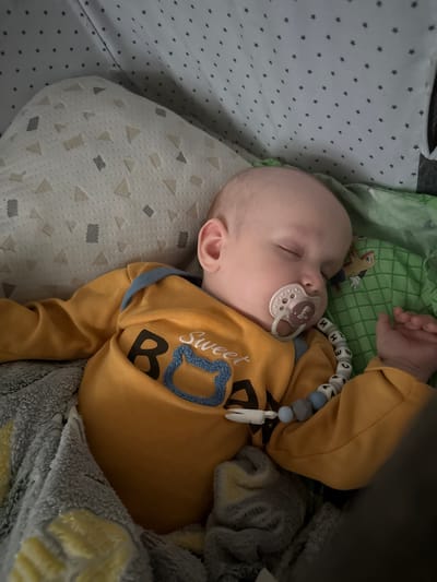 A baby sleeps in a crib, wearing a yellow long-sleeved shirt with blue trim and the word "Sweet" embroidered on it, along with the letter "B" and a blue outline of a bear. The baby has a pacifier in its mouth, attached to a beaded string that spells out "BABY". The crib has patterned bedding with grey stars and a white geometric pattern. The background is out of focus, but appears to be part of a nursery. The image is a close-up, taken from above, and the lighting suggests it was taken indoors. The baby appears to be in the city of Бордуки, Russia, based on the context provided, though this cannot be visually confirmed from the image itself.